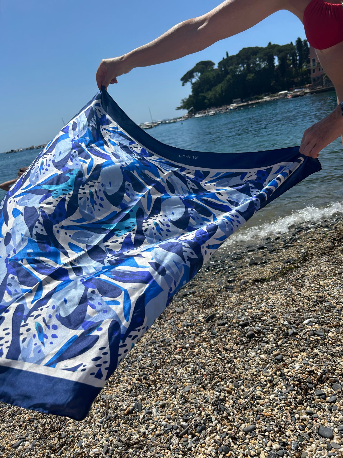 Beach shot of a blue and white Italian silk scarf blowing in the wind, designed in Lake Como, Italy. Ethically made using natural fibres, handwoven with a luxury finish