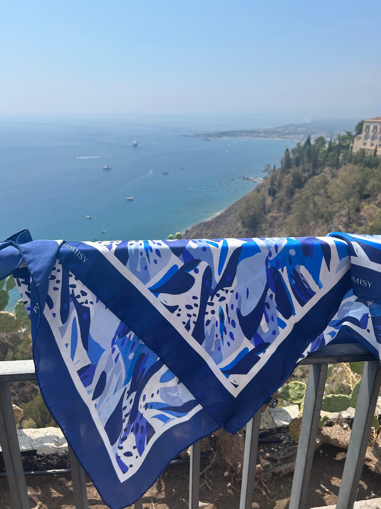 Blue and white Italian silk scarf ethically produced in Lake Como, Italy. The scarf is draped over a balcony with a view of the sea in the background, highlighting the Italian craftmanship of these silk scarves